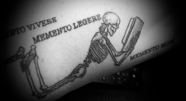 a black and white tattoo on a forearm of a skeleton laying forward, its feet kicked up behind and crossed at the ankles, propped up on its elbows and reading a book in its hands. 