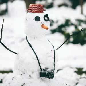 A snowman with a camera around his neck and an upside red pot on its head for a hat. 