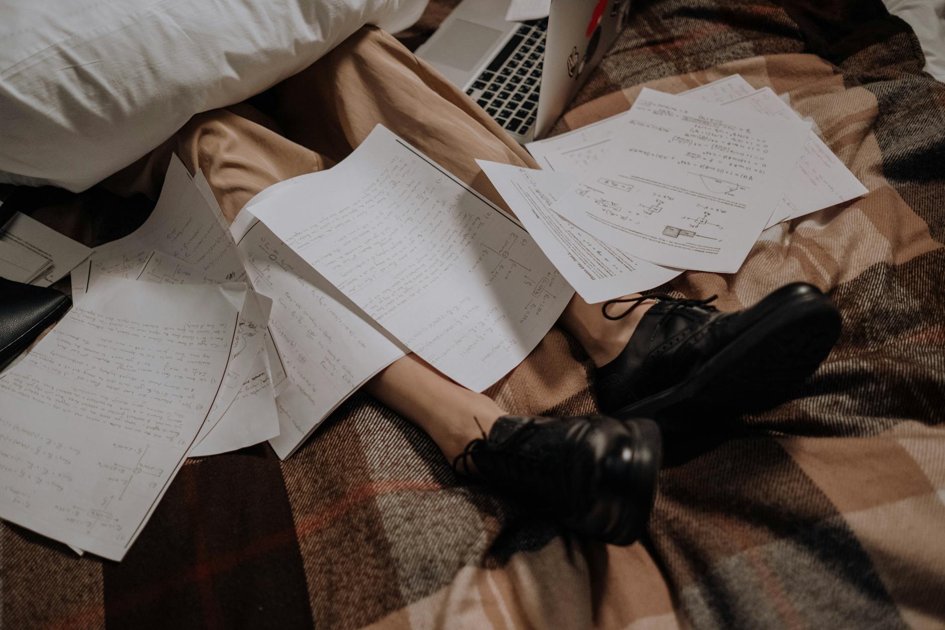 A person wearing tan pants and black shoes on a bed, with their feet up, with school papers scattered all around/over them and a laptop to the side.
